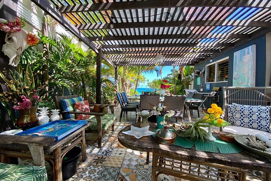 A lush, bohemian-style outdoor patio or deck with a slatted wooden pergola overhead. The floor is covered in colorful, patterned tiles. The space is furnished with rattan and wooden chairs, tropical plants, and decor like a decorative animal skull and ceramic pineapples. A clear view of the blue ocean is visible in the distance.