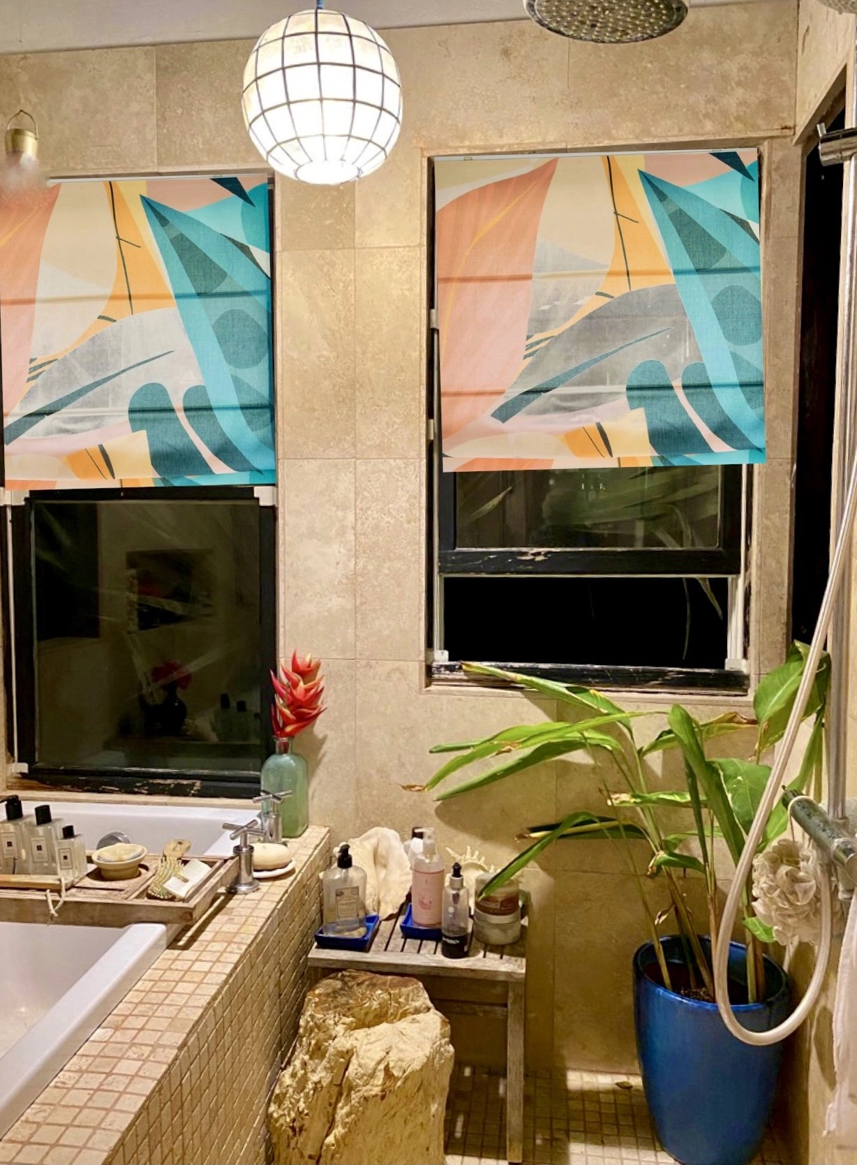 A bathroom with travertine tile walls and two windows covered with brightly patterned Roman shades featuring a tropical, colorful leaf design in teal, orange, and pink. A white bathtub is visible on the left, stocked with toiletries, and a potted green plant sits in a blue pot near a stone bench on the right.