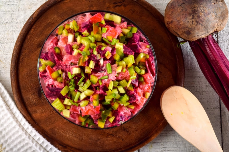 A vibrant overhead view of a bowl of diced salad, likely a variation of Olivier salad or Russian Vinaigrette. The salad is composed of diced pink and dark red beetroot mixed with a creamy dressing, topped with bright green diced celery and bell pepper. The bowl sits on a round wooden cutting board next to a wooden spoon and a whole, raw beetroot with its stem attached.