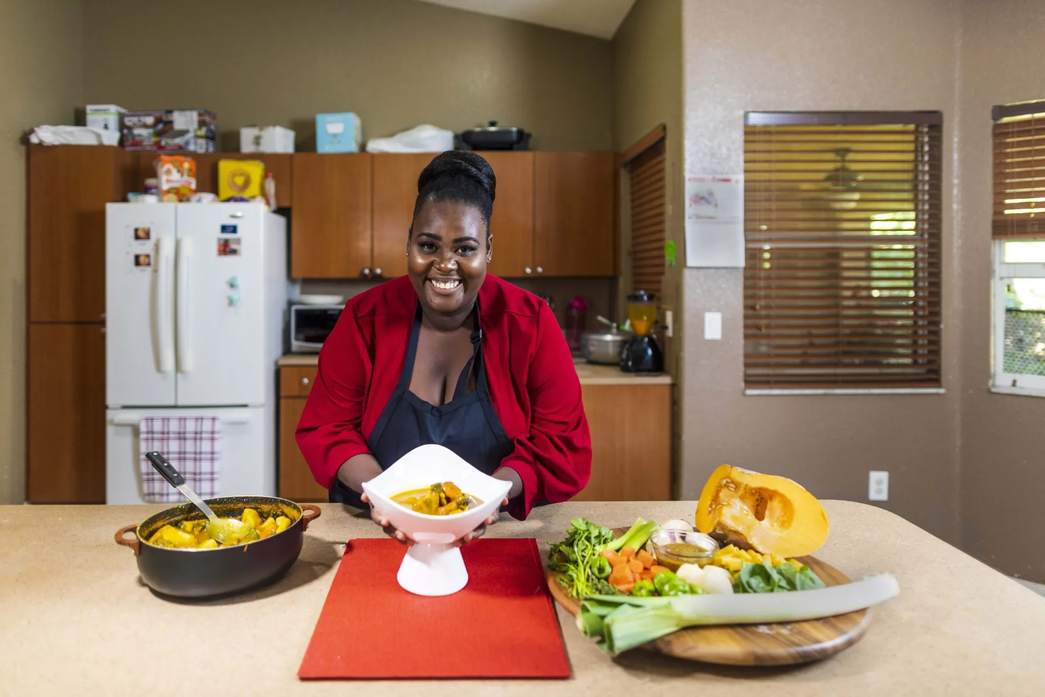 A smiling woman in a red jacket and black apron is standing behind a kitchen counter, holding up a white bowl containing a yellow curry or stew. A black pot of the same dish is on the counter to her left. To her right is a cutting board with a variety of fresh vegetables, including half a pumpkin or squash, leeks, carrots, celery, and microgreens.