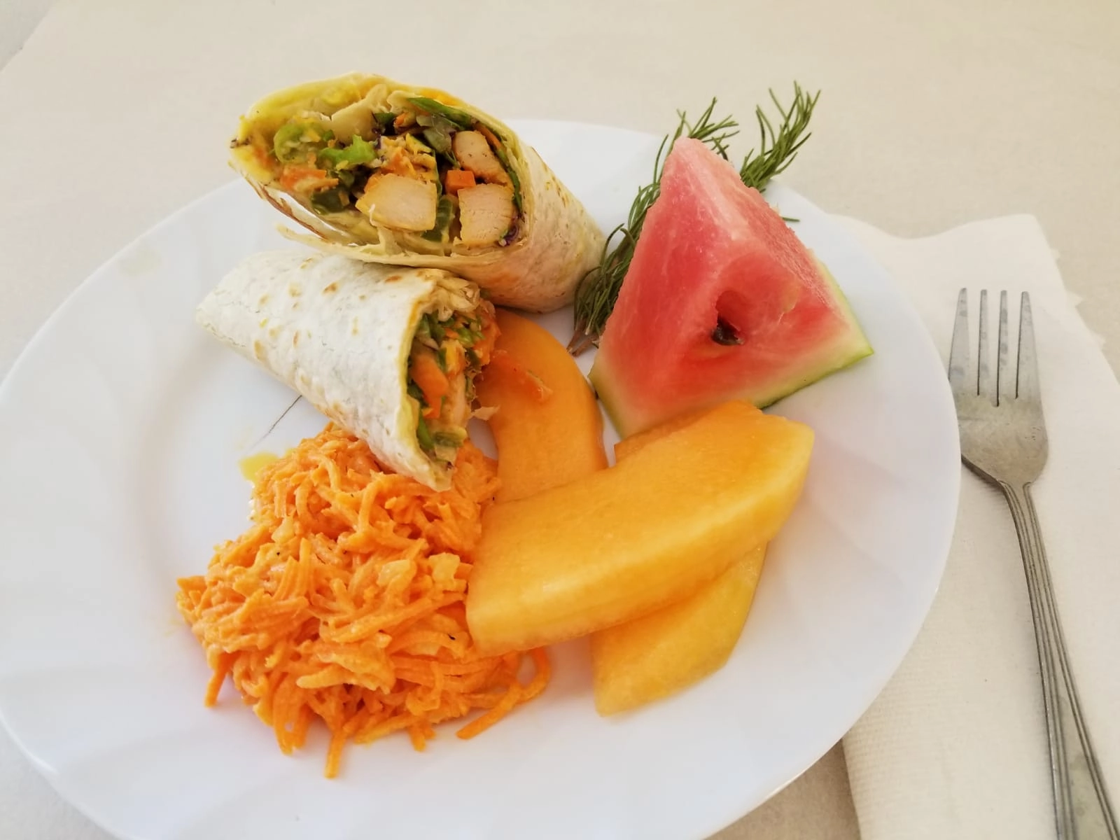 A white plate featuring a chicken and vegetable wrap cut in half, exposing the filling. The plate also holds a mound of shredded carrot salad (possibly with a sweet dressing), two slices of cantaloupe melon, and a wedge of pink watermelon garnished with a sprig of rosemary.
