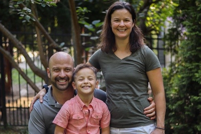 A candid, sunlit portrait of a smiling family of three outdoors, standing in front of dense green foliage and a dark metal fence. The father, a bald man, kneels on the left with his arm around his son. The son, a young boy with light brown hair, smiles brightly in the center, wearing a coral/pink collared shirt. The mother, a woman with dark hair, stands on the right wearing an olive green V-neck t-shirt and white pants.
