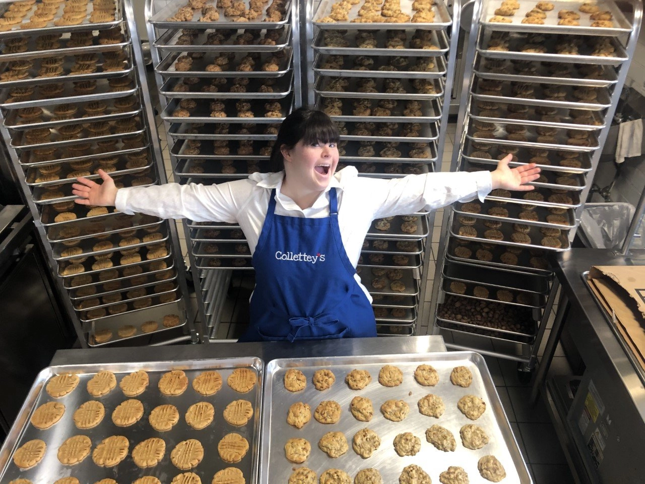 A young woman with Down syndrome, wearing a white shirt and a blue apron embroidered with "Collettey's," stands in a commercial kitchen with her arms spread wide and a joyful expression. In front of her are two trays of freshly baked cookies (one peanut butter, one oatmeal/drop cookie), and behind her are floor-to-ceiling racks filled with dozens of additional trays of baked cookies.