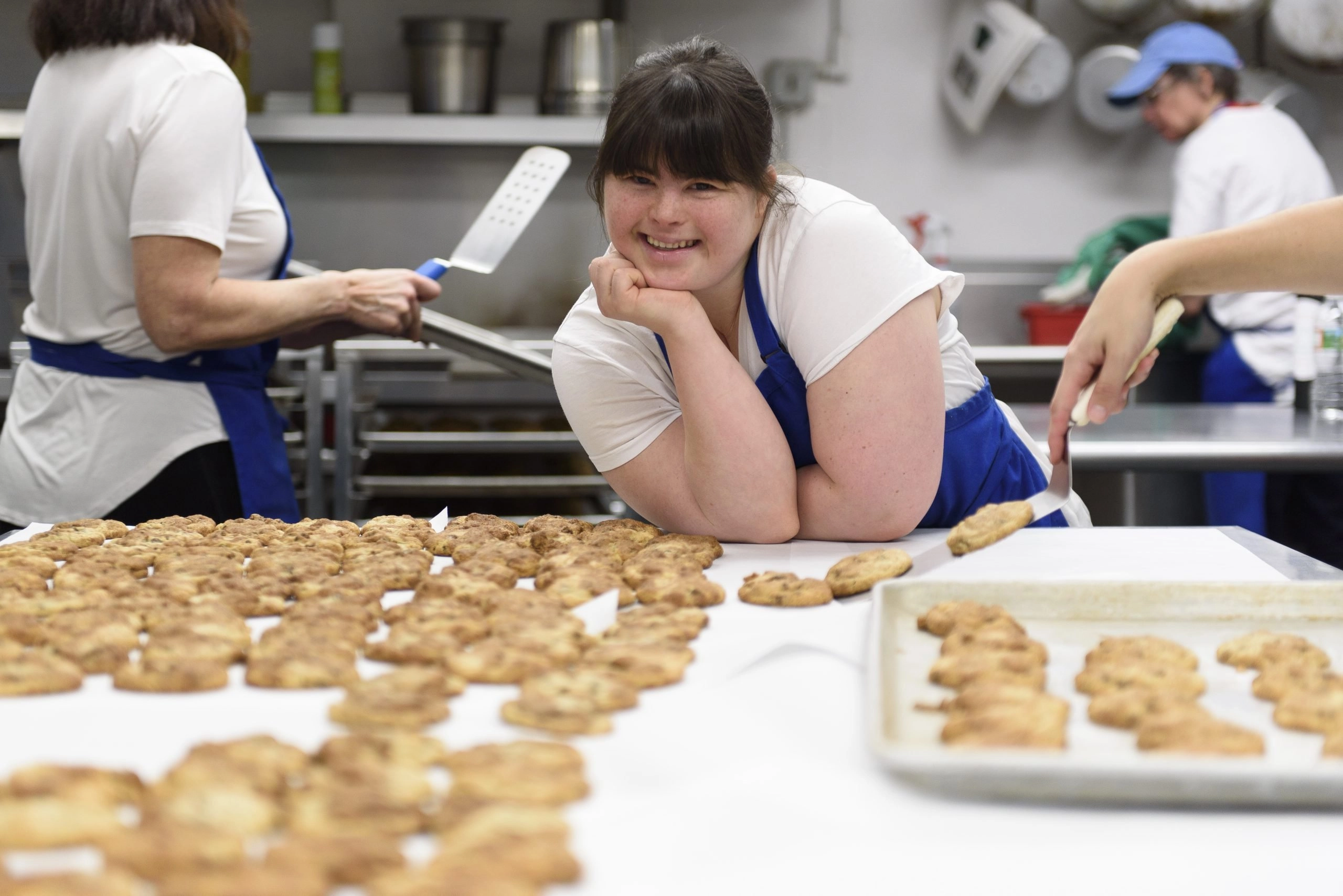 A close-up photo of a young woman with Down syndrome, wearing an apron, leaning on a counter and smiling brightly at the camera in a commercial kitchen. She is surrounded by large quantities of freshly baked oatmeal-style cookies, both on the counter and on a nearby baking sheet. Other workers are visible, blurred in the background.
