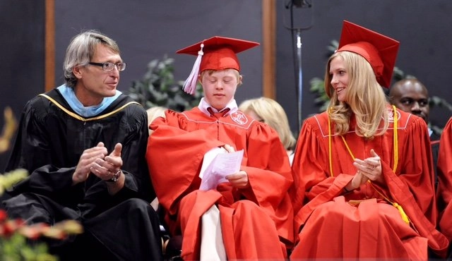 A photo taken at a graduation ceremony. A young man with Down syndrome is seated in the center, wearing a red graduation gown and cap, holding a paper. He is flanked by a man in a black and blue academic gown on the left and a young woman in a red graduation gown on the right. All three are smiling and clapping.
