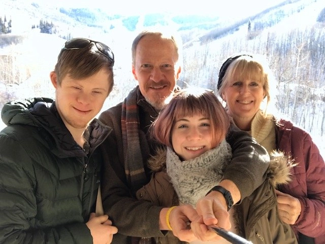 A selfie photo of a family of four, dressed in winter jackets and scarves, standing outdoors against a snowy, mountainous backdrop. The family includes a boy on the left with Down syndrome, a man, and a young woman and a woman on the right.