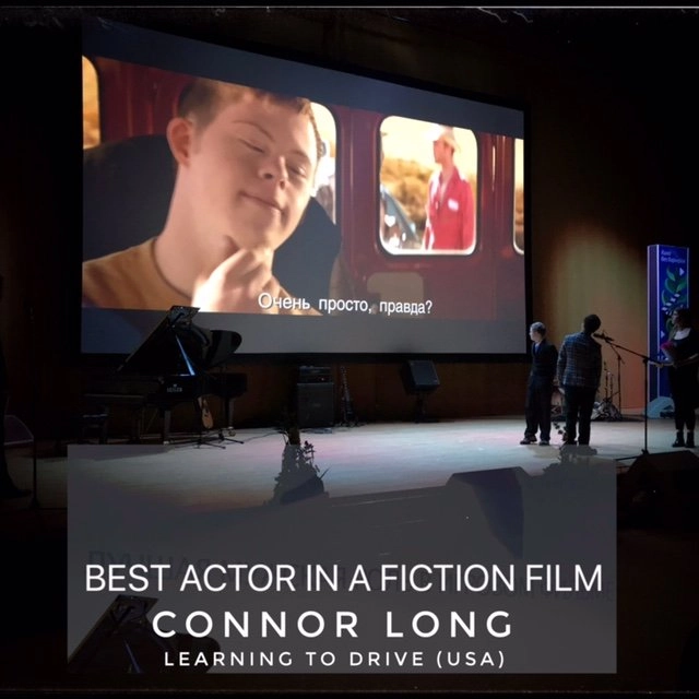 A large projection screen shows a still from a film, featuring a close-up of a young man with Down syndrome smiling. Russian subtitles are visible. Below the screen on a stage, a grand piano, speakers, and a small group of people are visible. A large overlay text block at the bottom reads: BEST ACTOR IN A FICTION FILM / CONNOR LONG / LEARNING TO DRIVE (USA).