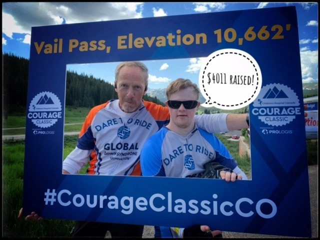 Two men, both wearing cycling jerseys, stand together and smile while holding a large, blue promotional frame for the "Courage Classic CO" event. The background shows a mountain landscape. Text on the frame reads "Vail Pass, Elevation 10,662'" and a speech bubble graphic says "$4011 RAISED!". One of the men is wearing sunglasses and a Down syndrome awareness logo on his shirt.