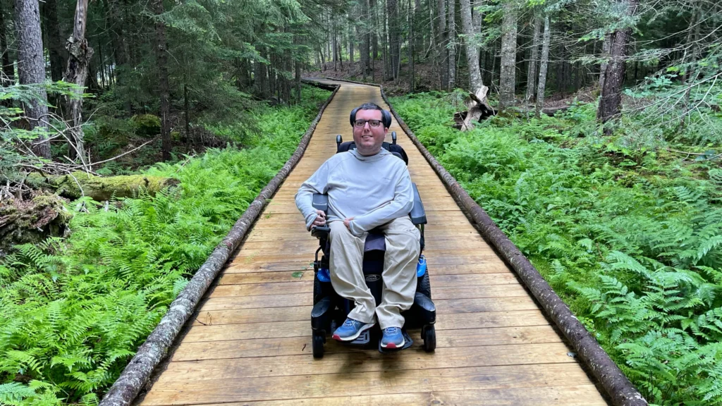 A man in casual clothes smiles from his power wheelchair, which is on a raised wooden boardwalk that's bordered in logs and runs through a lush forest.