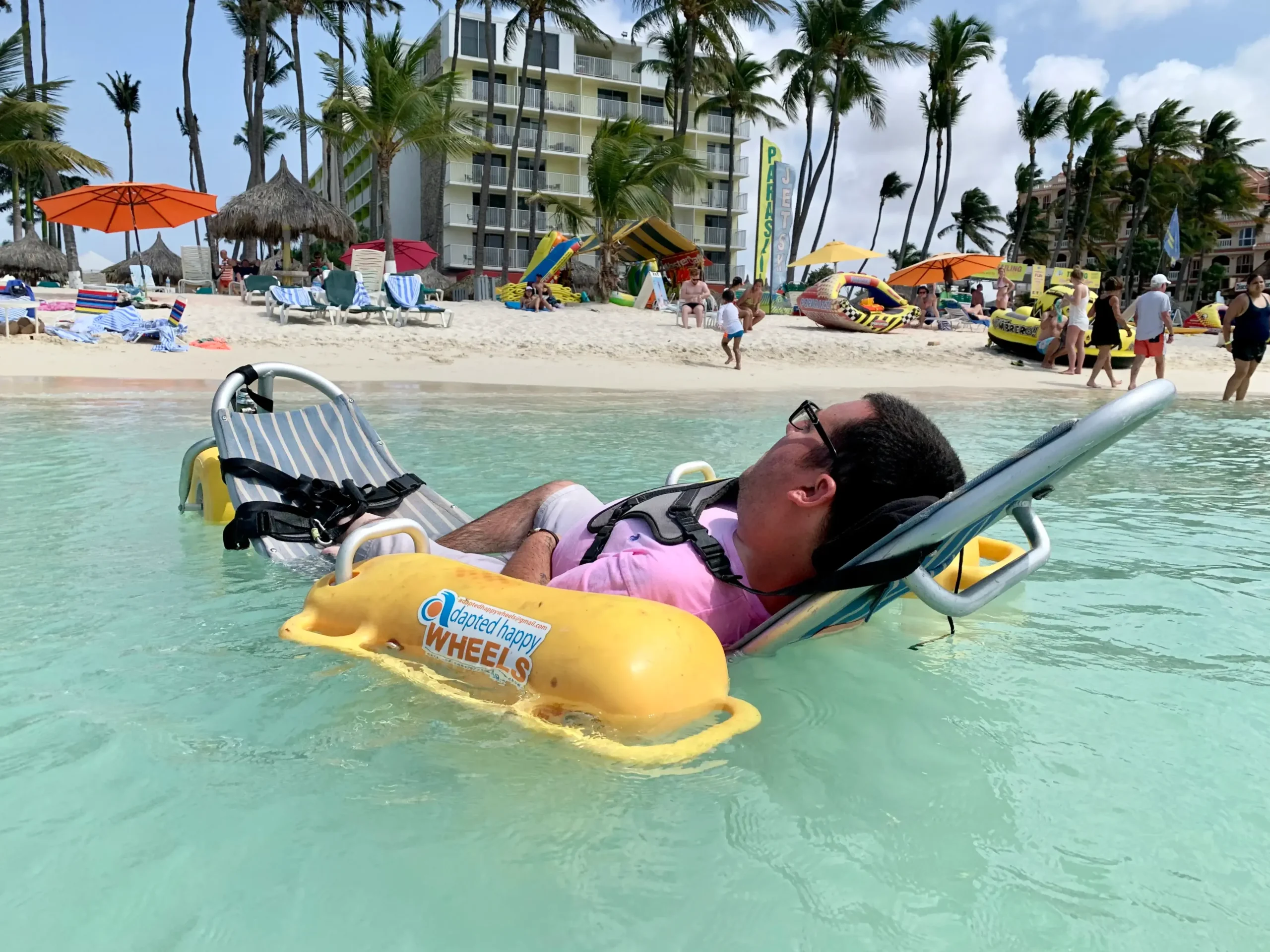 A man is floating on his back in clear, shallow turquoise water using an assistive floating chair. The chair has a striped blue and white fabric seat and is stabilized by yellow pontoons labeled 'adapted happy wheels'. He is wearing a pink shirt, white shorts, and a life vest. In the background, a sunny, white sand beach is visible, featuring palm trees, a large resort hotel, and beachgoers enjoying the water and sun under orange umbrellas.