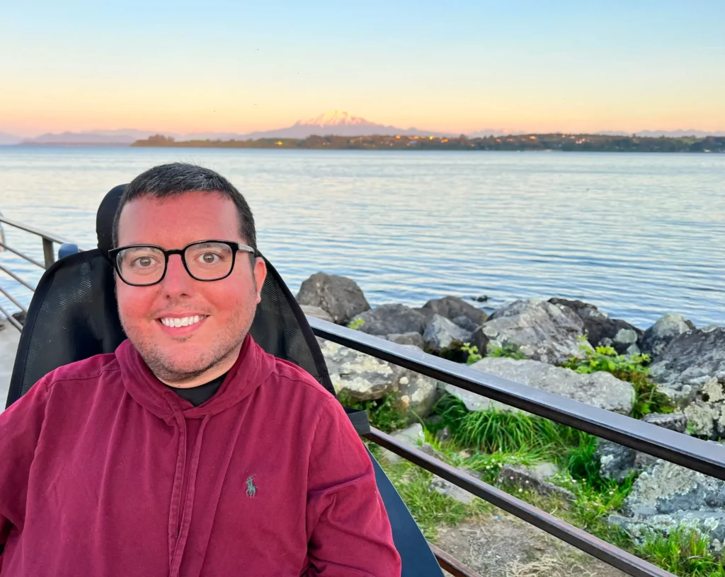 A smiling man with glasses, wearing a maroon hoodie, sits outdoors in what appears to be a wheelchair. He is positioned by a railing overlooking a calm lake or large body of water. In the background, there are rocks and green foliage along the shore, and a large mountain with a snow-capped or light-colored peak is visible in the distance beneath a pale orange and blue sunset sky.