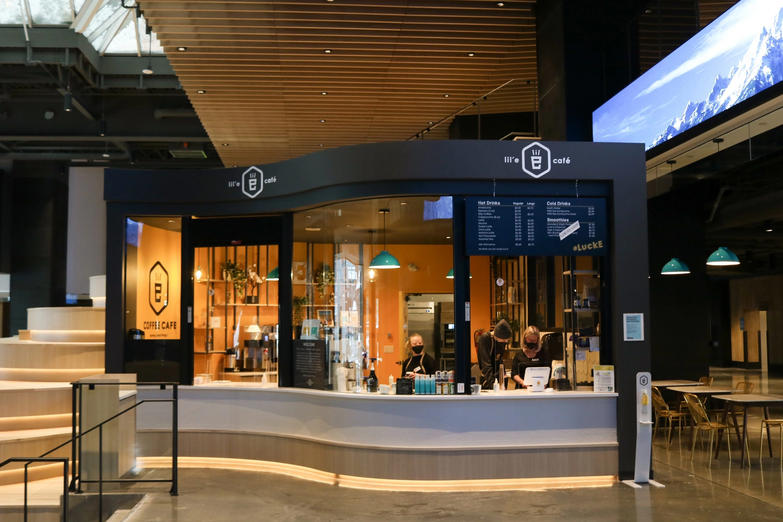 An exterior view of a modern coffee shop counter named "lil' E cafe" and "COFFEE CAFE" with a black and wood finish. The interior walls are bright orange. Two staff members wearing masks are visible behind the white counter. A digital menu board hangs on the right, listing hot drinks, cold drinks, and smoothies. The cafe is located in a spacious indoor atrium with wooden steps, gold tables and chairs, and a large screen displaying mountain scenery in the background.