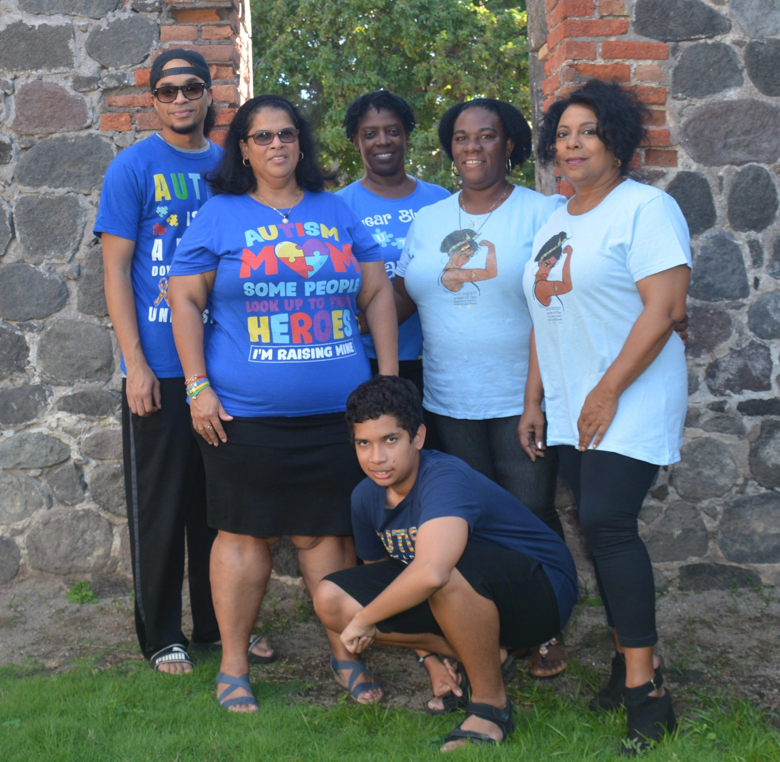 A group photo of five women and one young man standing in front of a stone and brick wall, possibly the same structure as in the previous image. The women are wearing various blue or light blue t-shirts, including one that reads "AUTISM MOM: SOME PEOPLE LOOK UP TO HEROES, I'M RAISING MINE." The young man in the foreground is crouching. All appear to be posing together for a group shot.