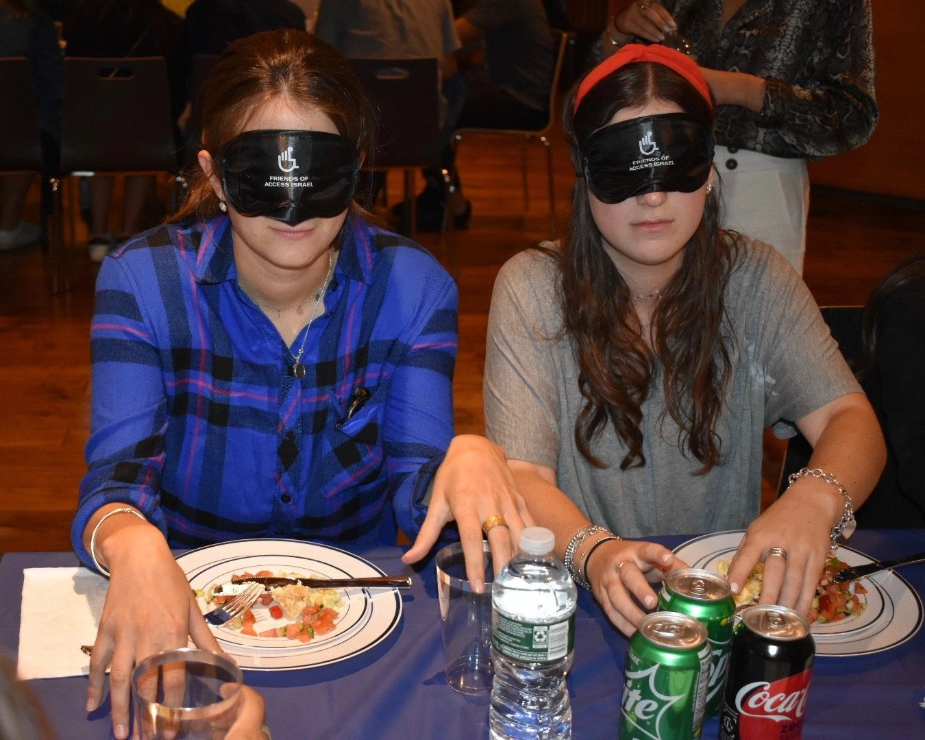 Two young women seated at a table covered with a blue cloth, both wearing black blindfolds over their eyes with a logo for "Friends of Access Israel." They are participating in a meal, with plates of partially eaten food, forks, a water bottle, and soda cans visible. The woman on the left is wearing a blue plaid shirt, and the woman on the right is wearing a gray shirt and a red headband.