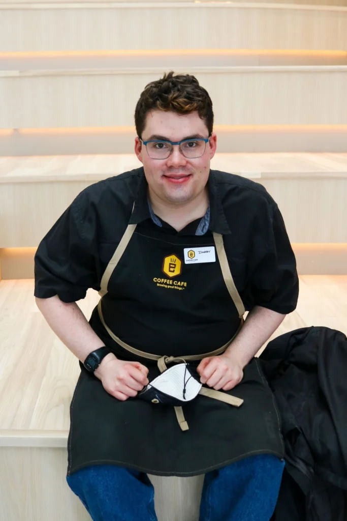 A young man with dark hair and glasses, wearing a black shirt and a black apron with the "Coffee Cafe" logo, sits smiling on modern wooden steps with embedded lighting. He is holding a white cloth or face mask in his hands. His name tag, visible on his apron, reads "Timothy."