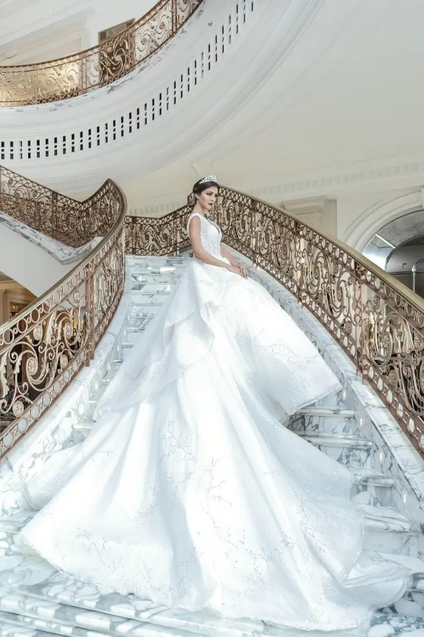 A woman in a white, off-the-shoulder wedding gown with a long, sweeping train that features subtle embroidery. She is standing on a grand, curving staircase made of white marble with intricate gold railings, looking over her shoulder.