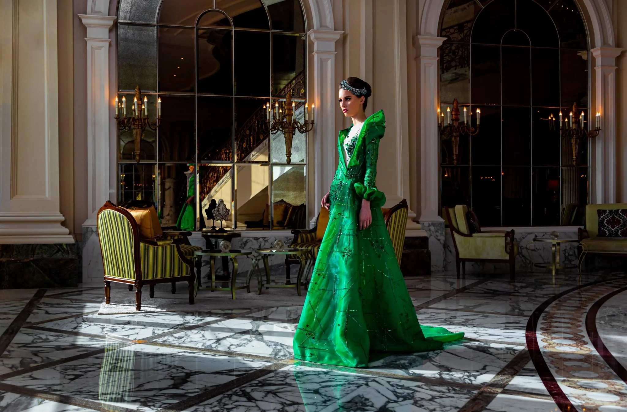 A woman standing in the center of a luxurious hall, wearing a long, dramatic, high-collared emerald green gown with puffed sleeves. The hall features a polished black and white marble floor, tall columns, and large arched windows reflecting the ornate interior and chandeliers.