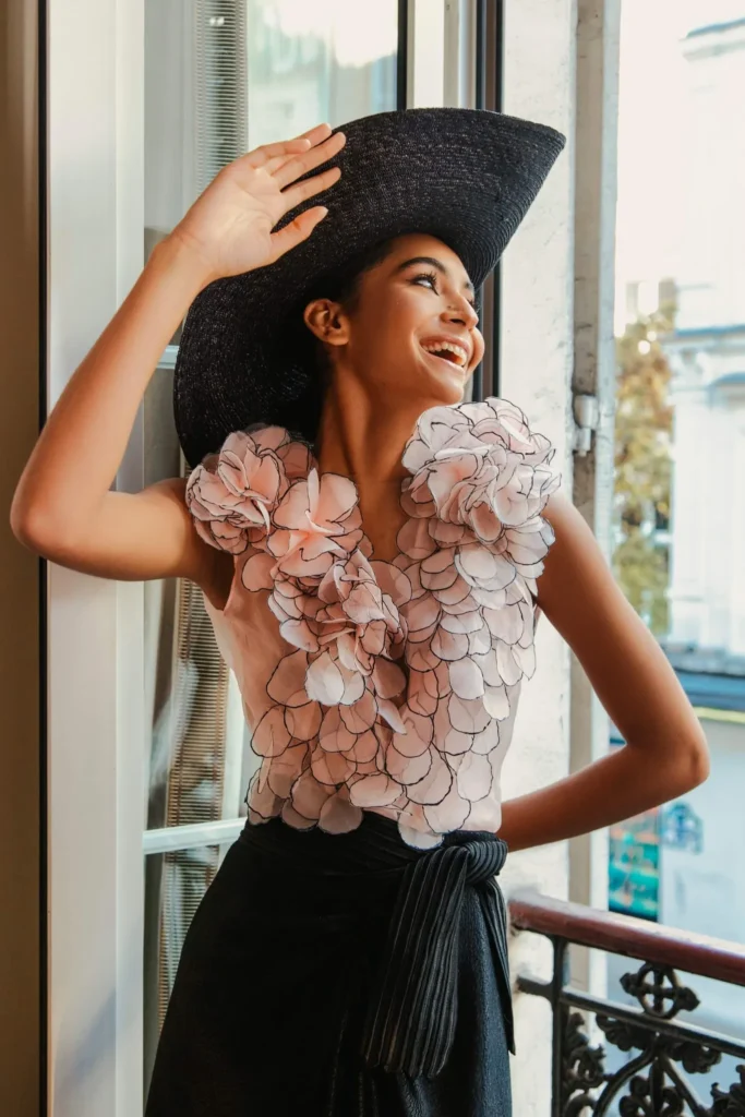 A portrait of a smiling woman standing on a balcony by a window, wearing a large, black woven sun hat and a pale pink top decorated with large, textured floral appliqués around the shoulders and neckline. Her other hand rests on her hip over a dark, belted skirt.