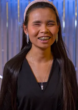 A portrait of a young woman with long, straight dark hair and braces, wearing a black top and a pink headband. She is smiling, has her eyes closed, and is wearing long, glittering silver drop earrings and a silver pendant necklace. She is standing in front of a blurred, shimmering blue and white background.
