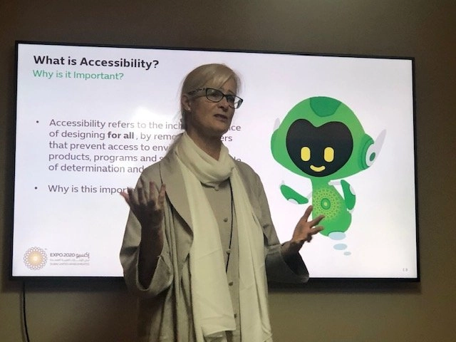 A middle-aged woman with blonde hair and glasses, wearing a light trench coat and scarf, stands in front of a large screen giving a presentation. The slide is titled "What is Accessibility? Why is it Important?" and features an image of a small, friendly green robot mascot on the right.