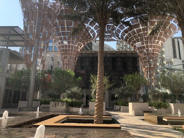 A modern outdoor plaza with several water features, small fountains, and low planters containing greenery. A prominent palm tree stands in the center. In the background, there is a large, distinctive building with a lattice canopy structure extending over the outdoor space, designed to look like giant, inverted umbrellas or funnels.