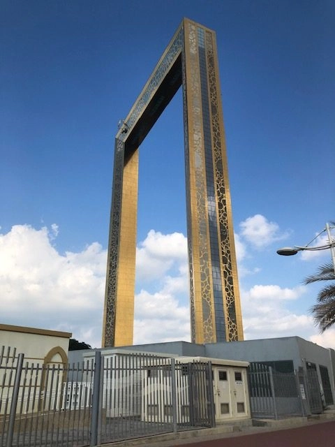 The iconic Dubai Frame, a tall, rectangular, golden-clad structure, stands against a bright blue sky with white clouds. The structure sits atop a low, grey service building surrounded by a metal fence.