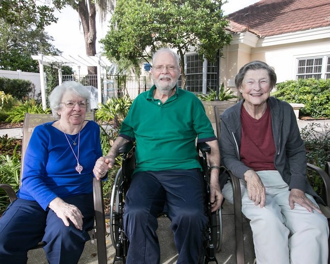 Three elderly people are sitting outdoors in patio chairs, smiling at the camera. The man in the center is wearing a green polo shirt and dark pants, seated in a wheelchair. A woman in a royal blue shirt and pants is seated on his left, holding his hand. Another woman in a dark grey hoodie and light pants is seated on his right. They are in a well-maintained garden area outside a building, possibly an assisted living community.