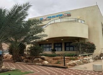 The exterior of a modern, tan-colored building, possibly a visitor center, situated in a desert landscape. The building has a curved second-story balcony with signage in Hebrew. Date palm trees and rocky landscaping are visible in the foreground.