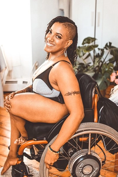 A portrait of a smiling Black woman with a shaved undercut and long dreadlocks, sitting in a wheelchair. She is wearing a black and gray bralette/lingerie top and is barefoot, looking towards the camera. She has tattoos on her arm and leg, and is wearing beaded bracelets. The background shows a light-filled indoor space with a wooden floor and plants.
