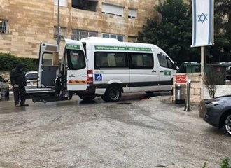 A white paratransit van with a wheelchair symbol and green lettering is parked on a ramp, facing right, with its rear lift lowered. A person in a dark jacket is standing near the lift, and an Israeli flag on a pole is visible on the right. The background includes a beige apartment building.
