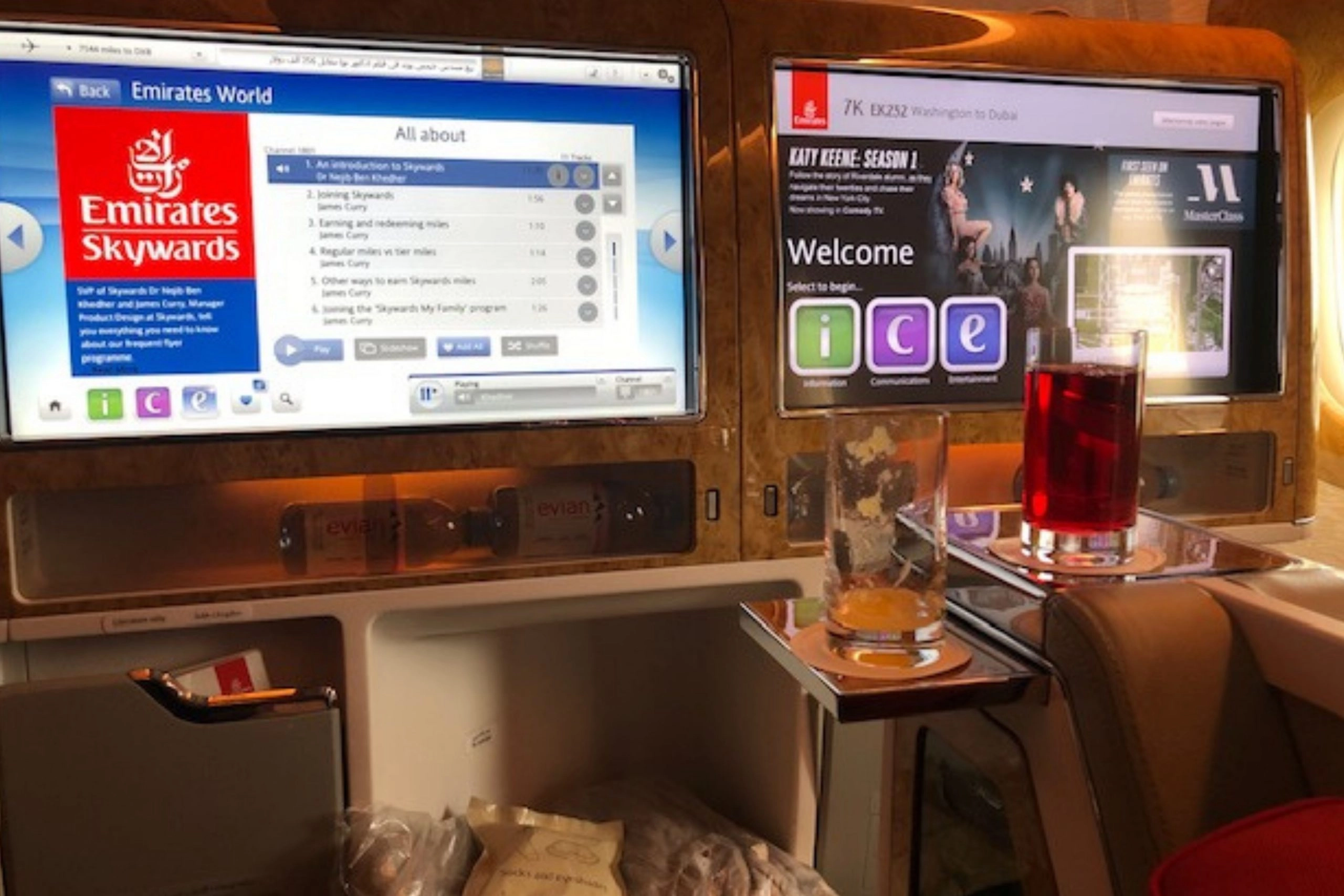 A view of a business or first-class suite on an Emirates airplane, showing two large, wood-paneled in-flight entertainment screens. The left screen displays the 'Emirates Skywards' loyalty program, and the right screen displays the 'ice' welcome page for movies and entertainment. A glass of red drink and an empty glass sit on the side table.