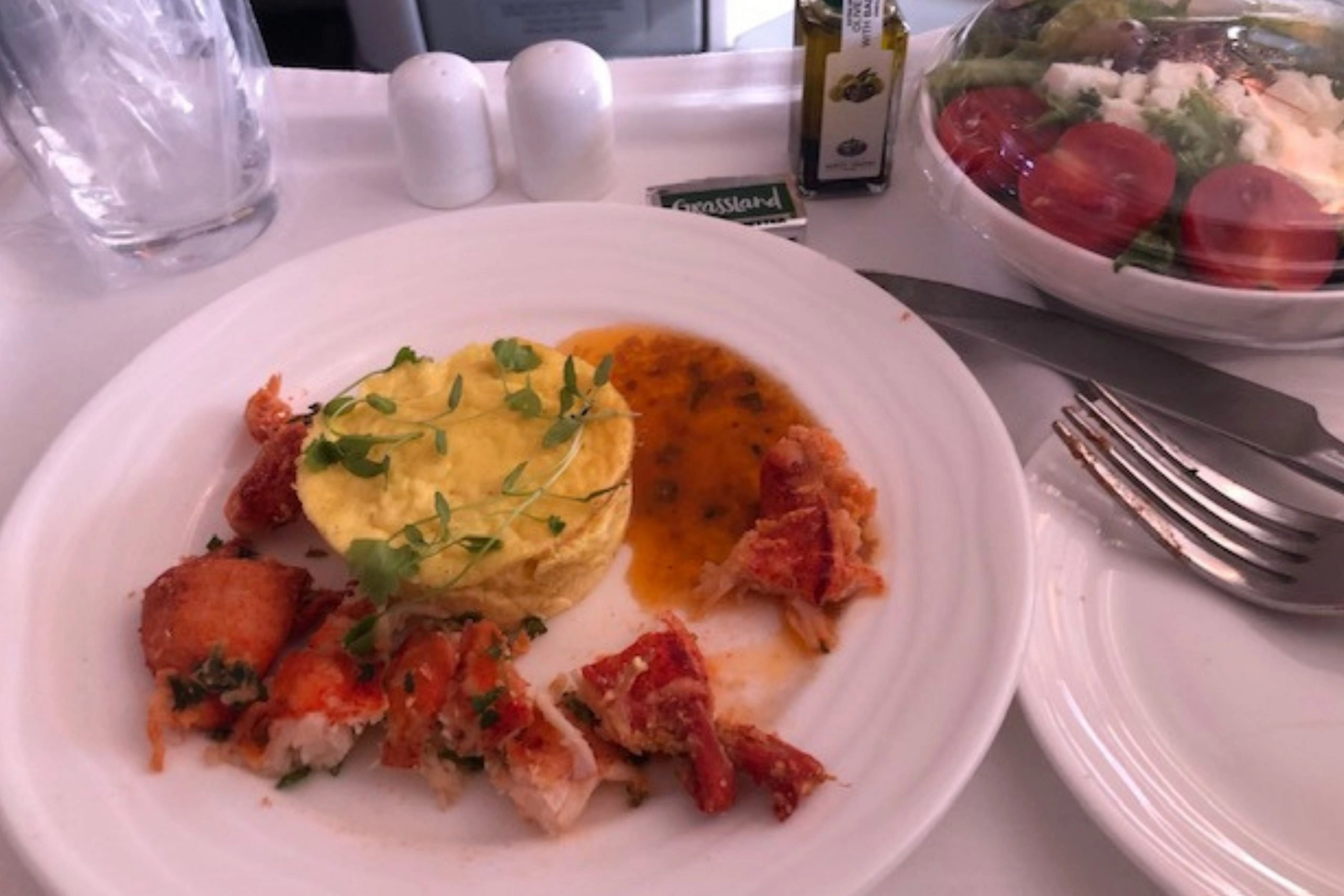 A close-up of a premium airplane meal served on a white plate. The main dish features chunks of lobster and a mound of mashed potatoes or polenta with an orange sauce and microgreens. A side salad in a plastic container, salt and pepper shakers, and a small bottle of olive oil are also visible on the tray table.