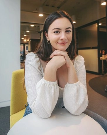 A portrait of a young woman with long, dark hair, wearing a white blouse with puffed sleeves, sitting at a white round table in an office or lounge setting. She is smiling gently and resting her chin on her clasped hands. A yellow accent chair is partially visible, and the background is blurred, showing glass dividers and other office furniture.