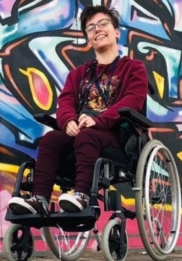 A young person with short, dark hair and glasses is seated in a modern manual wheelchair, smiling brightly up and slightly away from the camera. They are wearing a maroon hoodie, matching maroon pants, and sneakers. The background is a brightly colored wall covered in vibrant graffiti art.