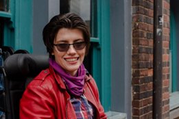 A woman in a red leather jacket, purple scarf and sunglasses smiles in front of grey and green window sashes and a brick wall.