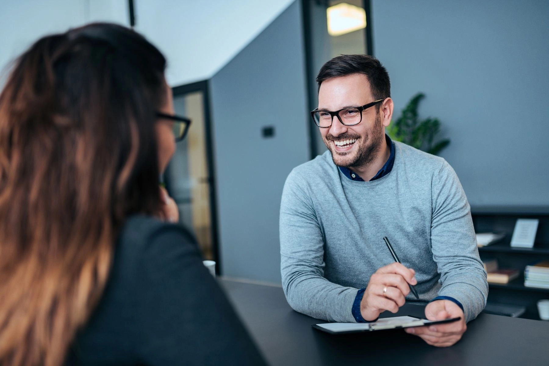 A professional photo of a man and a woman sitting across a desk from each other in a modern office setting, likely during an interview or meeting. The man, wearing a gray sweater over a collared shirt and black-rimmed glasses, is smiling broadly while holding a pen and clipboard. The woman, whose back is to the camera, has long brown hair and is also wearing glasses. The background is a mix of blue-gray and white walls.