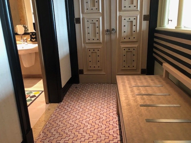 The entryway to a hotel room showing a pink and white patterned tile floor leading to a set of heavy, pale wooden closet doors with dark metal studs. A wooden bench with metal accents runs along the wall to the right, and an open doorway on the left leads to the bathroom.