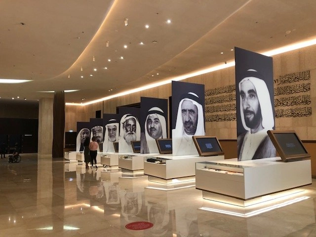The interior of a modern museum or exhibition hall with polished marble floors and curved ceilings. A row of large display panels featuring black and white portraits of the founding Sheikhs of the UAE is visible. In the foreground, there are interactive white display plinths.
