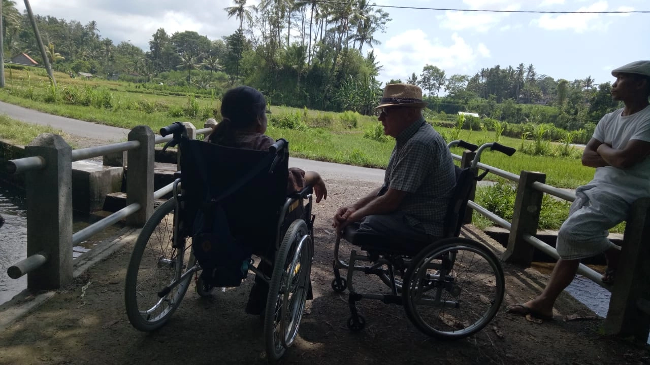 Two people in manual wheelchairs are facing each other and talking near a low concrete railing next to a small bridge or stream crossing in a lush, green, rural area. The background features rice paddies and palm trees. A third man wearing a light hat and white clothing stands on the far right, observing.