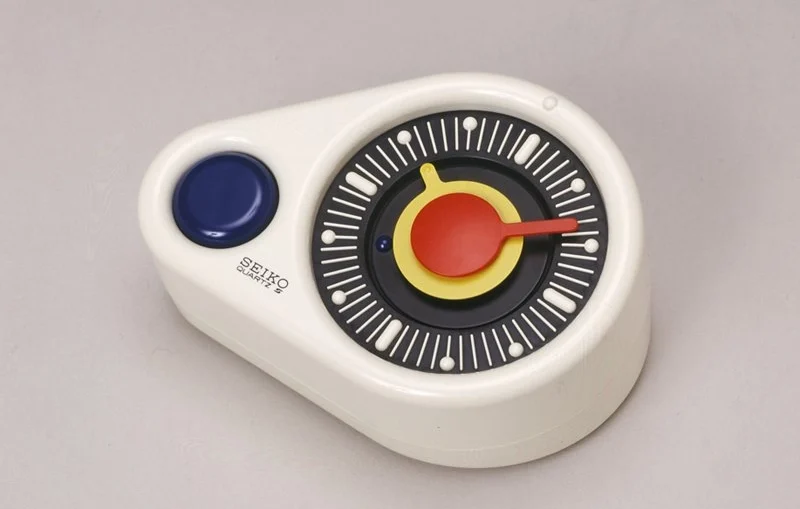 A close-up photograph of the "SEIKO QUARTZ Touch-me" table clock. The clock has a smooth white, organic-shaped body with a dark blue circular button on the left. The face features a black dial with white tactile markers, a central yellow disc, and red hour and minute hands designed for easy visibility and touch reading.