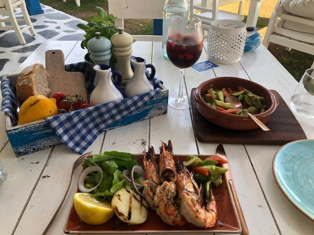 A delightful outdoor meal on a white plank table. A plate of large, grilled prawns seasoned with herbs, accompanied by a side of mixed greens and a grilled lemon half, is in the foreground. Other items include a bowl of roasted vegetables, a wine glass filled with a red drink, and a wooden basket containing bread, tomatoes, and a yellow fruit, all set on a blue and white checkered cloth.