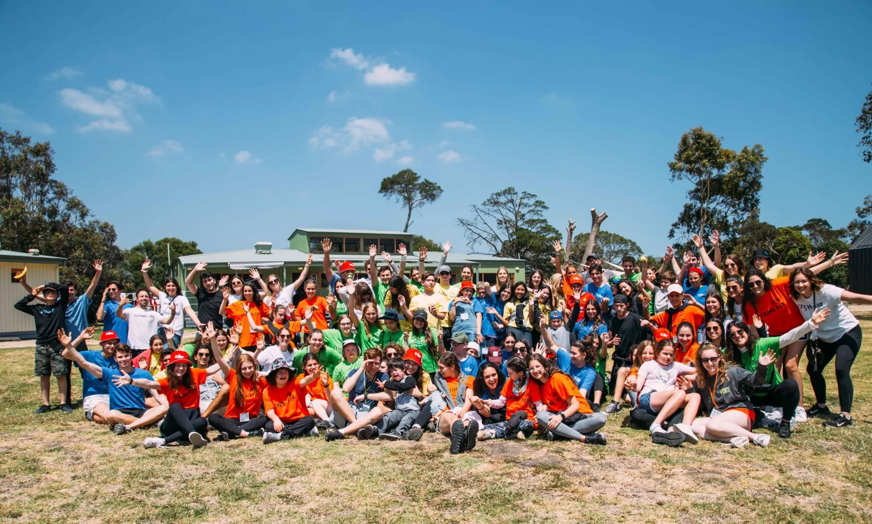 A large, diverse group of approximately 80 teenagers and young adults, many wearing brightly colored t-shirts in orange, blue, green, and red, sits and stands together on a grassy field with their hands raised in the air, smiling happily. They are outdoors on a sunny day with a light blue sky and white clouds, and a low, green-roofed building and trees are visible in the background.