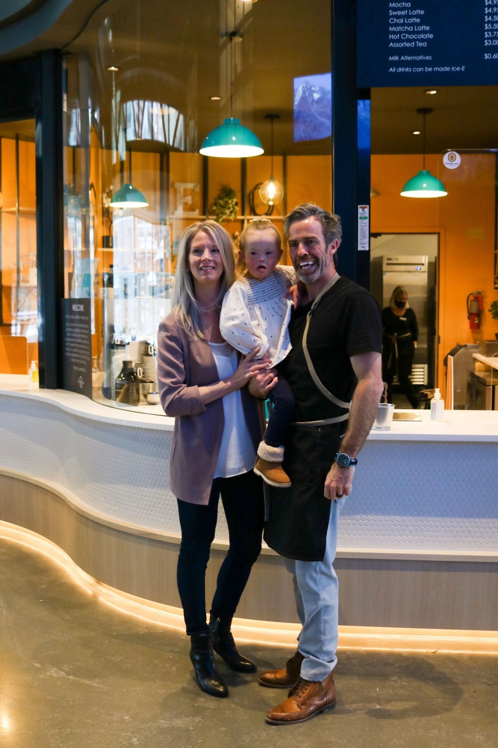 A full-length photo of a family standing in front of a modern coffee counter. The father, on the right, wears a black shirt, a black and tan apron, and holds a young girl who is wearing a white dress and brown booties. The mother, on the left, wears a mauve blazer and a white top. A menu board is visible on the right, listing coffee drinks.