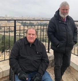 Two older men standing on an outdoor balcony or observation deck overlooking a cityscape, likely Jerusalem, under an overcast sky. The man in the foreground is seated in a wheelchair, wearing a black jacket and gloves, and the other man is standing next to him. The Dome of the Rock is visible in the distance over the railing.