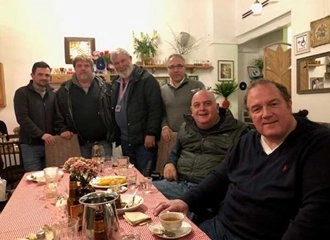 A group of six middle-aged to older men gathered around a table in what appears to be a cozy indoor restaurant. Three men are standing behind the table, and three men are seated in the foreground. The table is covered with a red and white checkered tablecloth and set with plates, drinks, and teacups.