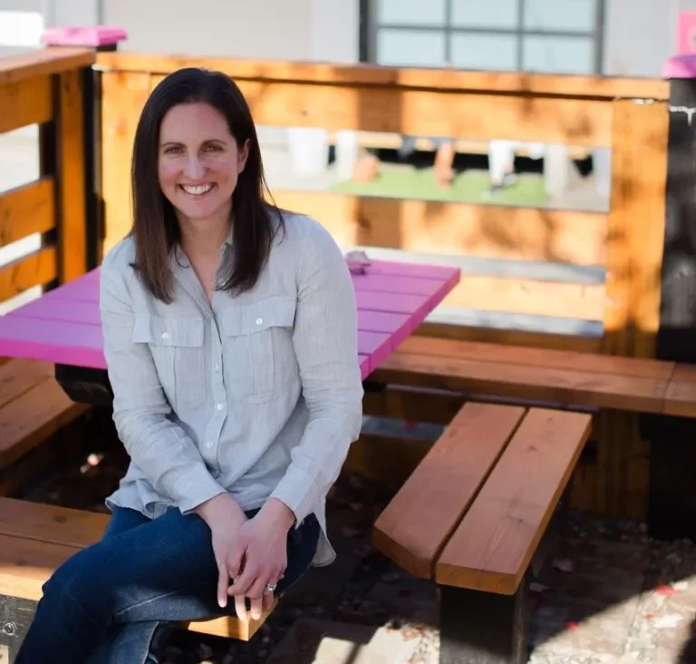 A headshot of Lisa Beecroft, the co-owner of Gabi & Jules, sitting on a wooden bench outdoors. She is wearing a light blue button-down shirt and jeans and is smiling at the camera. A bright pink table is visible behind her.
