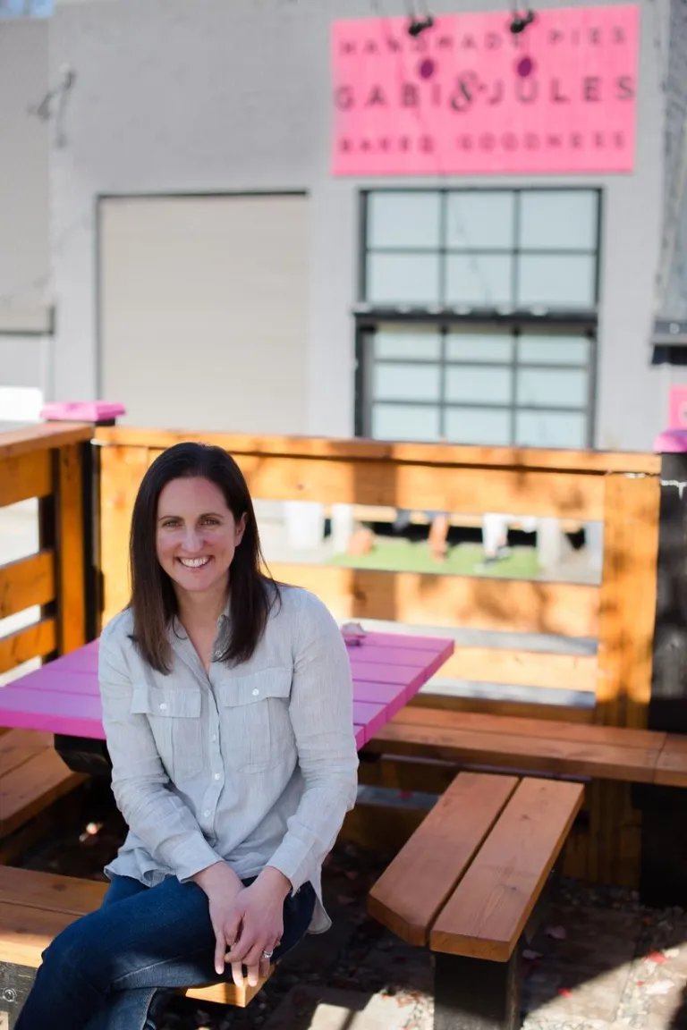 A headshot of Lisa Beecroft, the co-owner of Gabi & Jules, sitting on a wooden bench outdoors. She is wearing a light blue button-down shirt and jeans and is smiling at the camera. A bright pink table is visible behind her.