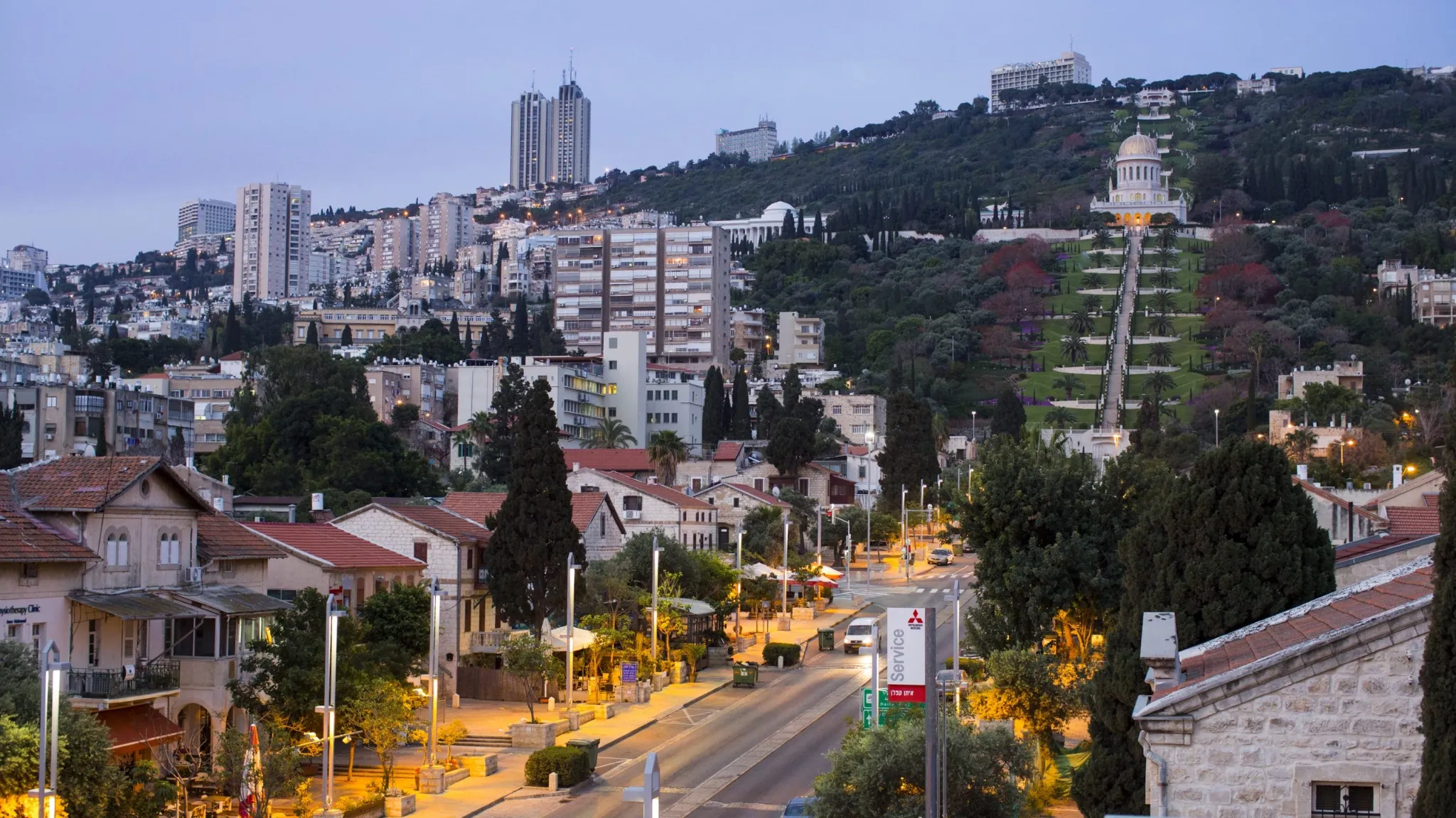 An evening or twilight view of the city of Haifa, Israel, showing a brightly lit street in the foreground with residential buildings. The Baháʼí Gardens and Shrine of the Báb, with its distinctive golden dome and terraced gardens, rise up the steep hillside in the middle ground, framed by modern high-rise buildings on the hillcrest.