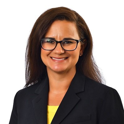 A woman with dark hair, wearing dark rimmed glasses and a business jacket, smiles at the camera.