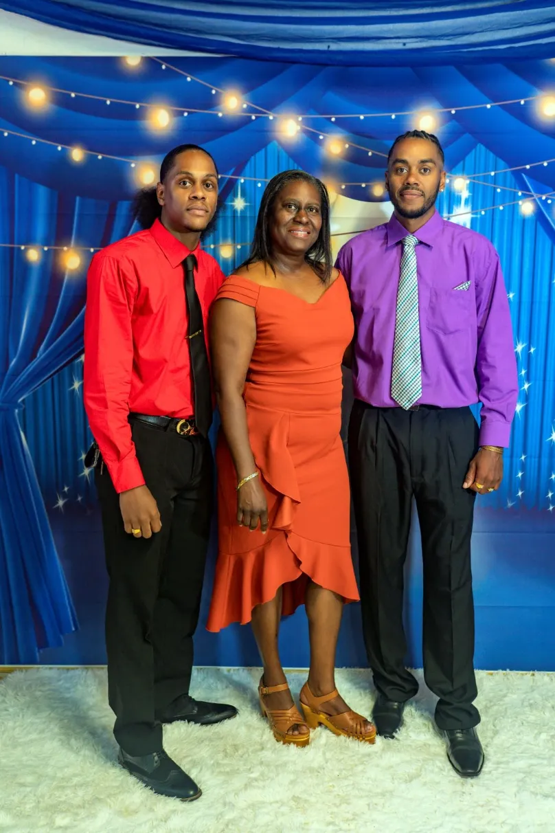 A formal indoor portrait of a woman and two young men standing in front of a blue background with white string lights. The woman in the center wears an orange, off-the-shoulder, ruffled dress with tan wedge sandals. The man on the left wears a red shirt and black tie, and the man on the right wears a purple shirt and a light grey patterned tie.